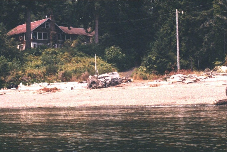 View of the old home from the water, beach and driftwood in foreground, house nestled in forest