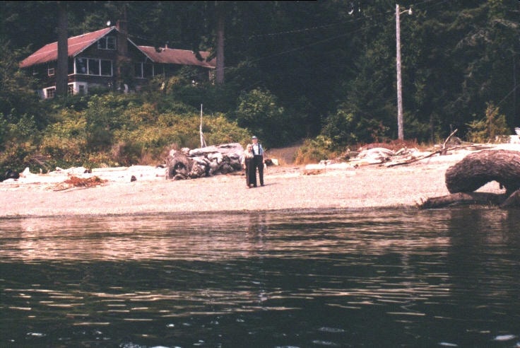 Mom and Dad standing on the beach in front of the old home, view from the water
