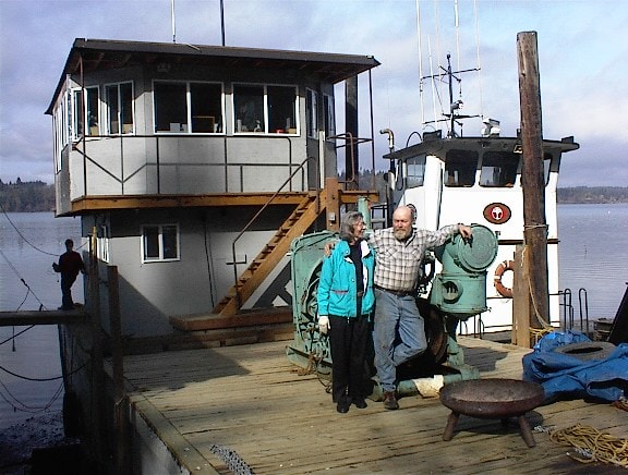 Two people standing on the dock in front of a tugboat and two-story boathouse