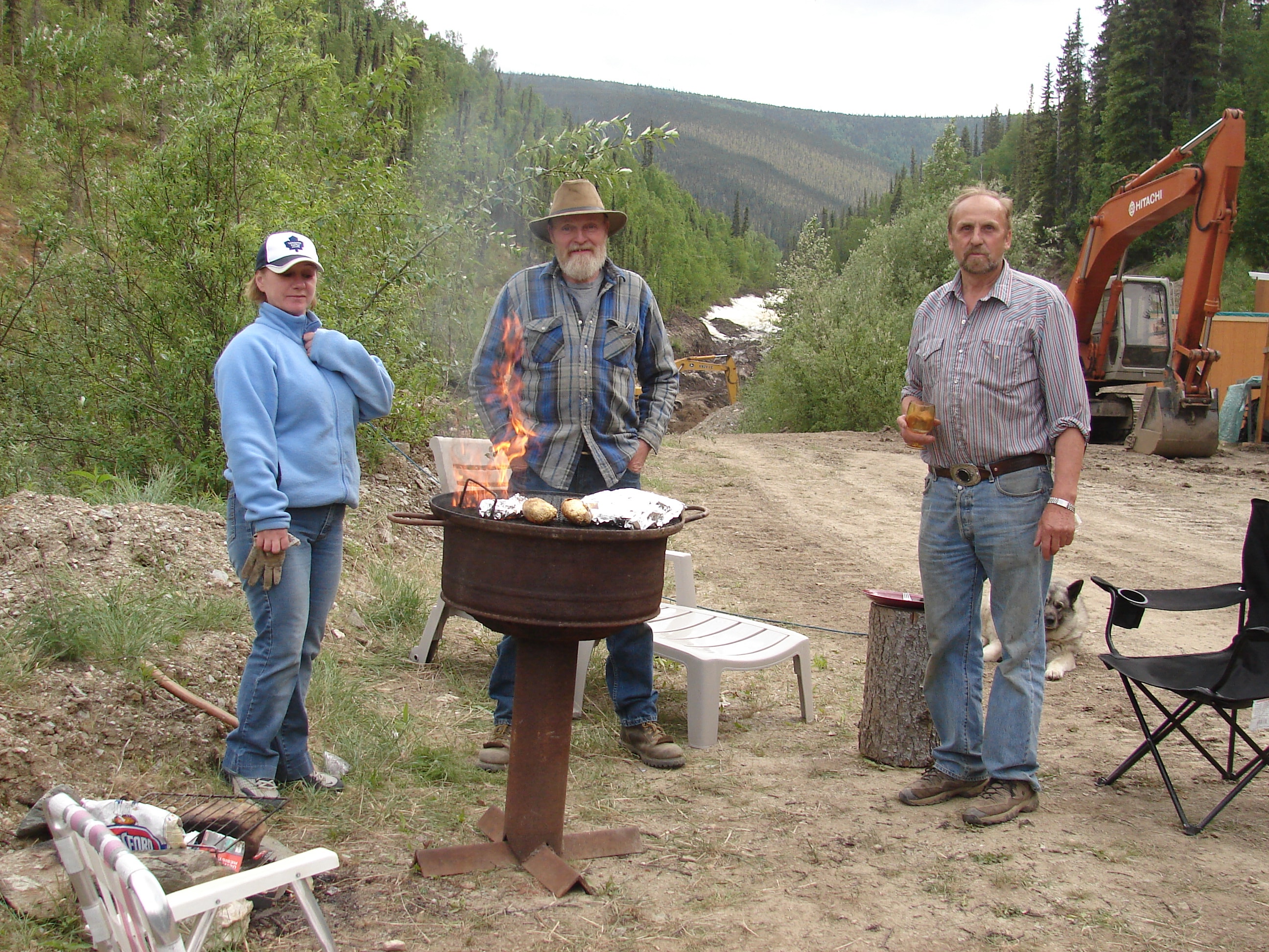 Three people at a campsite cookout with a fire pit and excavator in the background